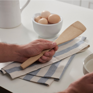 A person holds a left handed spatula while resting their hand on a Meema kitchen towel. A bowl of eggs is nearby on the counter top.