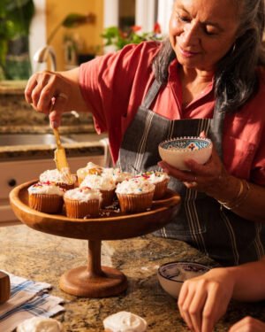 Woman in a kitchen decorating cupcakes on a wooden stand.