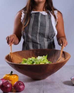 Person wearing an apron holding a extra large 20 inch wooden salad bowl with salad ingredients on a gray surface.